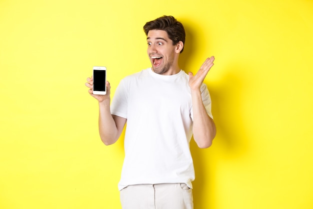 Image of amazed guy looking at mobile phone screen with surprised face, standing excited against yellow background