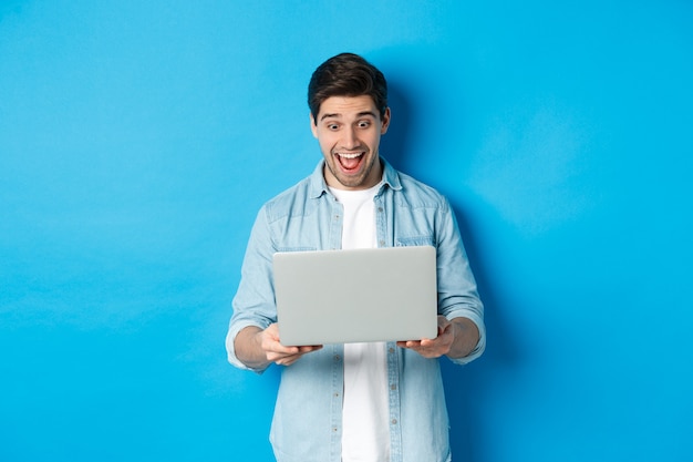 Image of amazed and happy man reacting to special offer in internet, looking at laptop excited, standing against blue background.