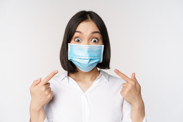 Image of amazed young asian woman pointing at herself while wearing medical face mask standing over white background