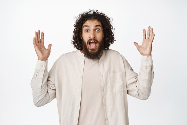 Image of amazed young man looking excited spread hands sideways and stare in awe big super cool news standing over white background