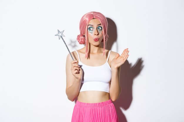 Image of amused cute girl with pink wig and bright makeup, dressed up as fairy for halloween party, holding magic wand and looking excited, standing.