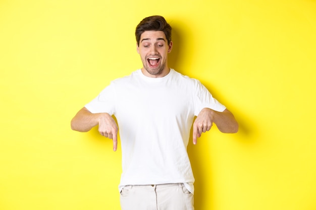 Image of amused handsome guy in white t-shirt, looking and pointing fingers down, standing over yellow background.