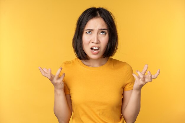 Image of angry asian woman shouting and cursing looking outraged furious face expression standing over yellow background