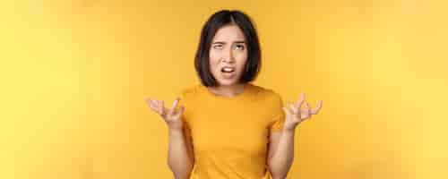 Free photo image of angry asian woman shouting and cursing looking outraged furious face expression standing over yellow background