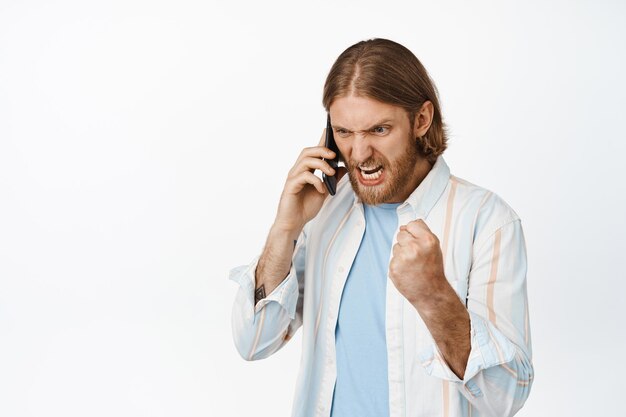 Image of angry blond bearded man cursing on phone call, screaming during conversation on mobile, shouting upset, standing against white background.