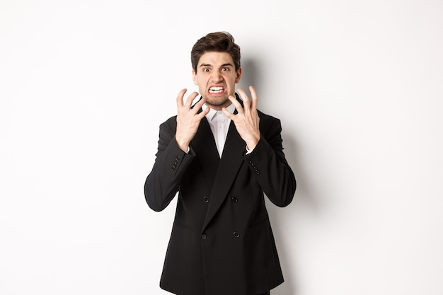 Image of angry businessman in suit, looking with furious expression and clenching fists, express hatred, standing mad over white background