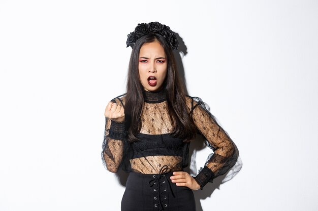 Image of angry and disappointed beautiful girl in halloween costume scolding someone. Girl shaking fist and looking with disapproval, wearing black gothic lace dress, standing white background.