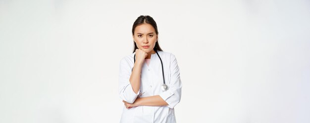 Image of angry nurse asian female doctor looking annoyed and bothered furrow eyebrows and sulking thoughtful standing over white background