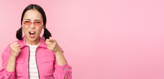 Image of angry pissed off korean adult female model shaking fists and shouting screaming outraged standing over pink background