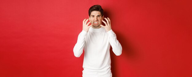 赤い背景に激怒して立って、怒りから顔をゆがめ、震えながら、白いセーターを着た怒り狂った男の画像。