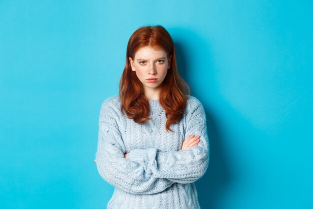 Image of angry redhead girl feeling offended, cross arms on chest and sulking, staring at camera mad, standing against blue background.