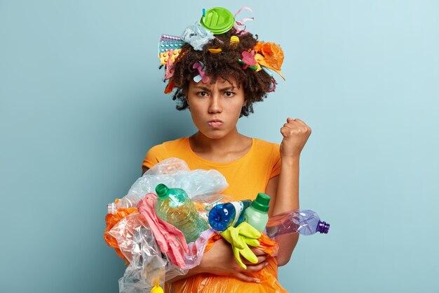 Image of annoyed black woman raises clenched fist, demands to be environmentally friendly, has grumpy facial exrpression, carries plastic waste, uses objects for recycling, stands over blue wall