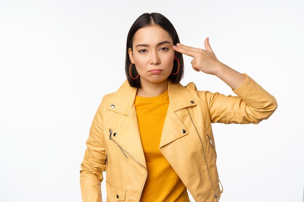 Image of annoyed and tired asian girl making handgun gesture fingers near temple looking bored and distressed standing against white background