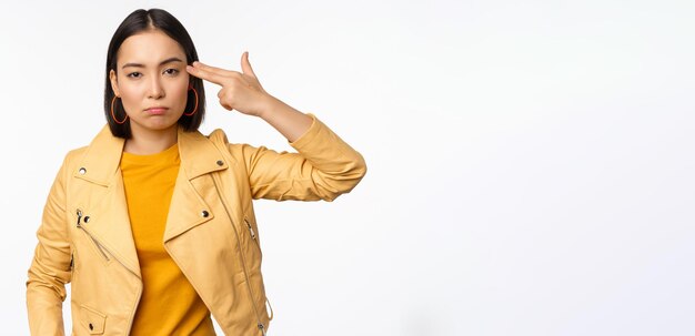 Image of annoyed and tired asian girl making handgun gesture fingers near temple looking bored and distressed standing against white background