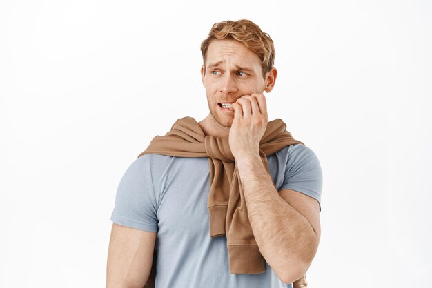 Image of anxious red head man biting finger nails and looking left with scared worried face staring at something with anxiety standing nervous against white background