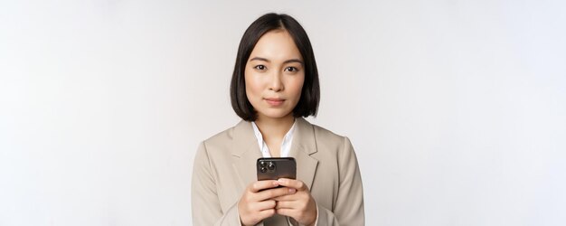 Image of asian businesswoman in suit holding mobile phone using smartphone app smiling at camera whi