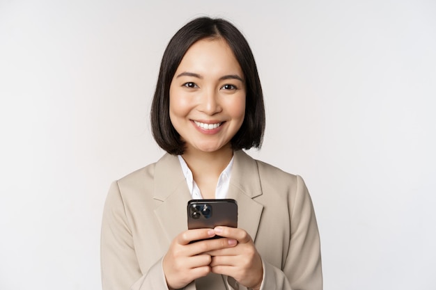 Image of asian businesswoman in suit holding mobile phone using smartphone app smiling at camera white background