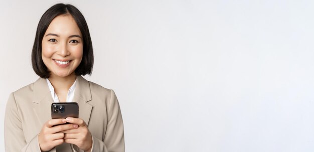 Image of asian businesswoman in suit holding mobile phone using smartphone app smiling at camera white background