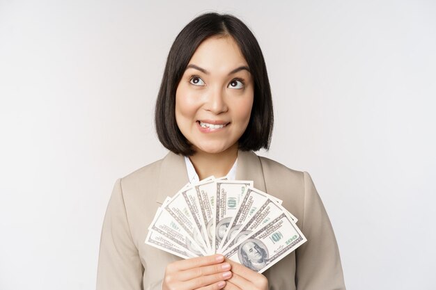 Image of asian corporate woman happy businesswoman showing money cash dollars and thinking standing in suit over white background