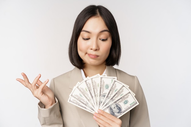 Image of asian corporate woman happy businesswoman showing money cash dollars and thinking standing in suit over white background