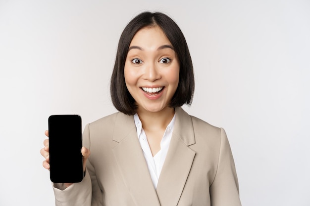 Image of asian corporate woman showing app interface mobile phone screen making surprised face expression wow standing over white background
