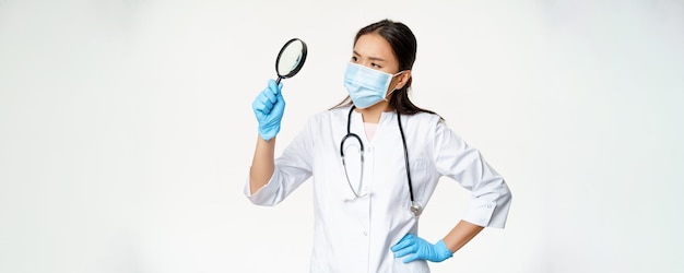 Изображение азиатской женщины-врача-ученого в медицинской маске и резиновых перчатках смотрит через увеличительное стекло в поисках чего-то на белом фоне
