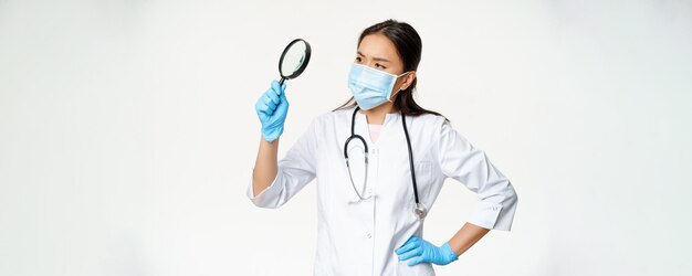 Image of asian female doctor scientist in medical mask and rubber gloves looks through magnifying glass searching for smth white background