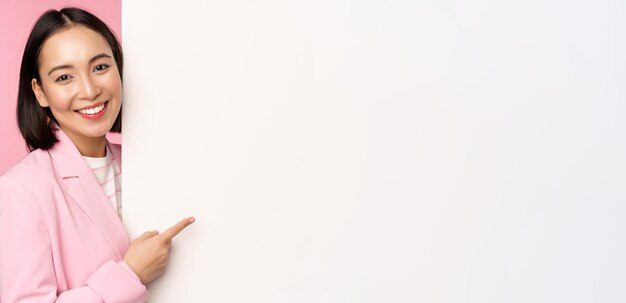 Image of asian female entrepreneur in suit pointing finger at board showing smth on white wall demonstrating chart or information standing over pink studio background