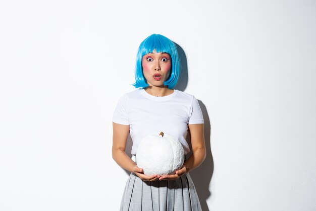 Image of asian girl in blue wig looking surprised and gasping, holding white pumpkin, standing.