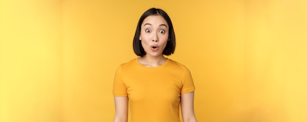 Image of asian girl looking surprised reacting amazed raising eyebrows impressed standing over yello