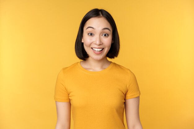 Image of asian girl looking surprised reacting amazed raising eyebrows impressed standing over yellow background