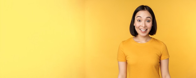 Image of asian girl looking surprised reacting amazed raising eyebrows impressed standing over yellow background