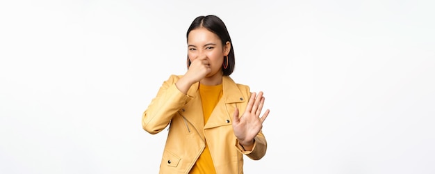 Image of asian girl shuts nose and reject smth with disgusting bad smell shuts nose standing over white background
