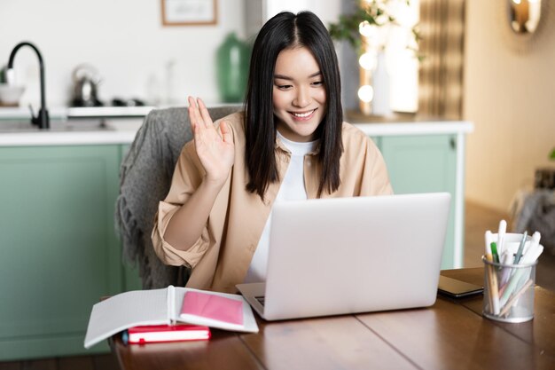 Image of asian girl on video call waving hand at her video chat on laptop and smiling student or tea...