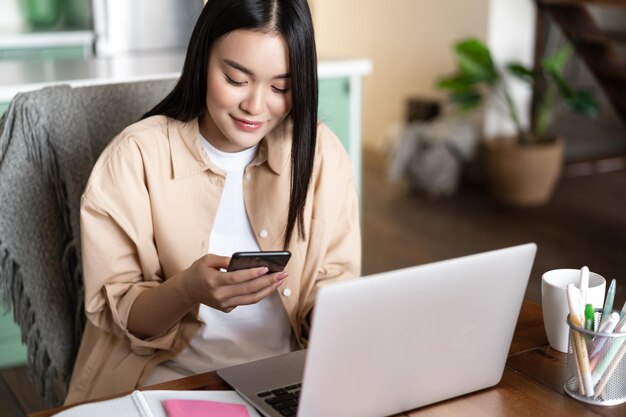 Image of asian woman checking phone messages working on laptop computer from home waiting for work c...