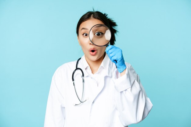 Image of asian woman doctor or nurse found smth looking through magnifying glass and stare surprised...