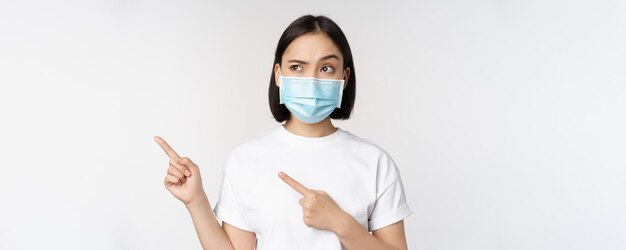 Image of asian woman in medical mask from covid looking confused left pointing at logo showing advertisement standing over white background