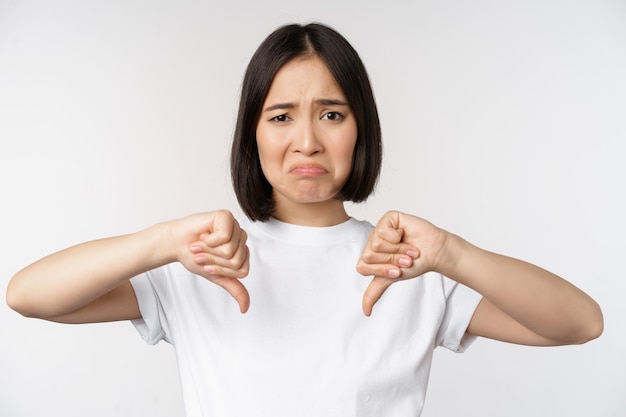 Image of asian woman showing thumbs down dislike smth looking disappointed standing over white background Copy space