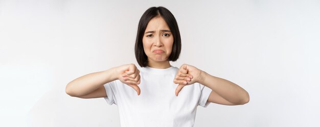 Free photo image of asian woman showing thumbs down dislike smth looking disappointed standing over white background copy space
