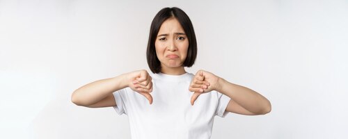 Image of asian woman showing thumbs down dislike smth looking disappointed standing over white background copy space