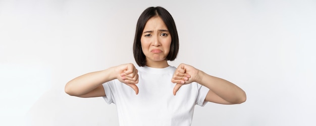 Image of asian woman showing thumbs down dislike smth looking disappointed standing over white background Copy space