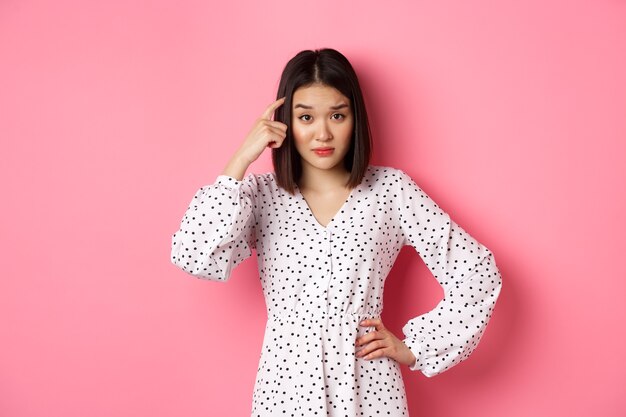 Image of attractive asian lady scolding person, pointing at head and staring at camera disappointed, are you stupid gesture, standing over pink background.