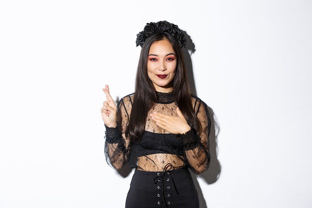 Изображение привлекательной азиатской женщины в платье партии Хэллоуина загадывая желание, держа руку на сердце и скрестив пальцы на удачу, стоя на белом фоне.