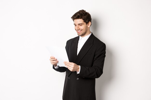 Image of attractive businessman in black suit, reading document and smiling, working on report, standing against white background