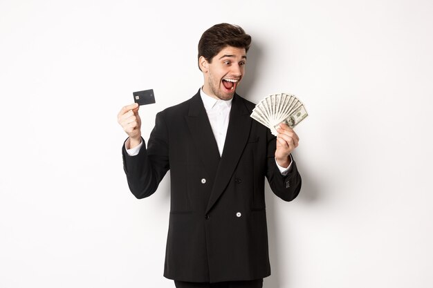 Image of attractive businessman in black suit, rejoicing, showing credit card and looking at money, standing against white background