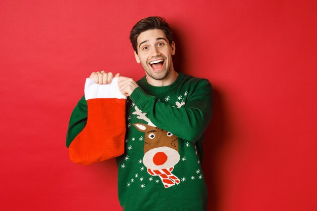 Image of attractive cheerful man in sweater, holding christmas stocking with gifts, celebrating winter holidays, standing over red background.