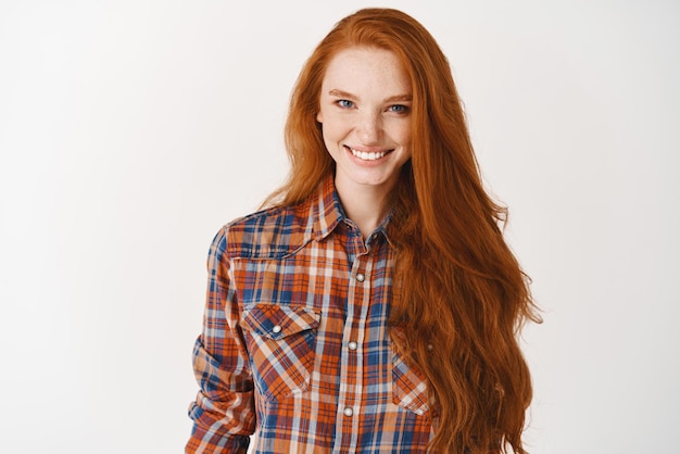 Изображение привлекательной женщины-модели с длинными здоровыми рыжими волосами, уверенно улыбающейся в камеру Молодая рыжая женщина, стоящая в клетчатой рубашке на белом фоне