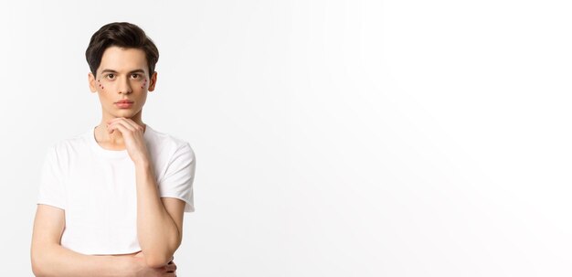 Image of attractive gay man in white tshirt having glitter on face and looking at camera serious standing over white background