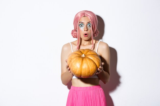 Image of attractive girl in pink wig looking surprised while holding pumpkin for halloween party, standing.