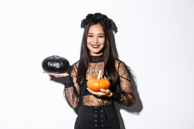 Image of attractive korean girl celebrating halloween in gothic lace dress, impersonating witch and holding two pumpkins.